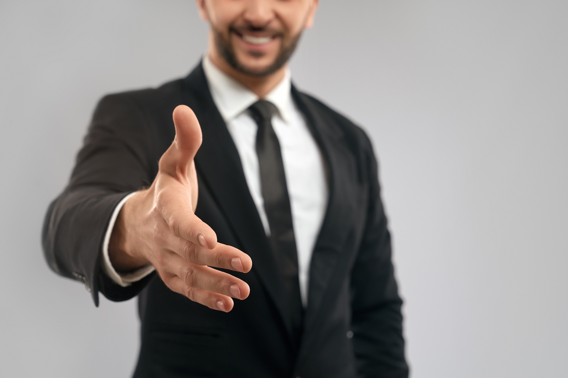 Close up of incognito smiling businessman in black formal suit giving hand for shake. Selective focus of friendly gesture, isolated on grey background. Concept of business, sealing deal.